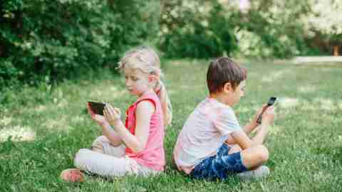 Boy and girl play games on smartphones outdoor. Kids digital gadget screen addiction. Children friends playing online sitting on grass in park. Contemporary problem of loneliness together.