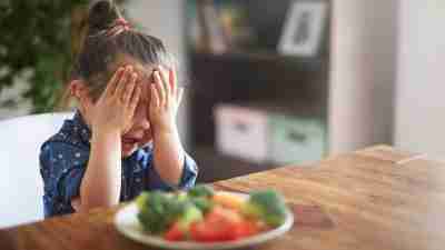 Girl with ADHD covers face with hand while sitting in front of plate of vegetables